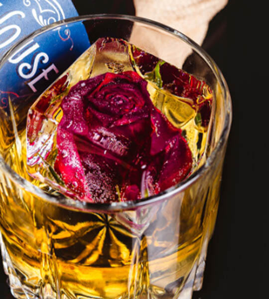 Glass containing golden liquor and a cube of Block Ice customized with a rose suspended within.